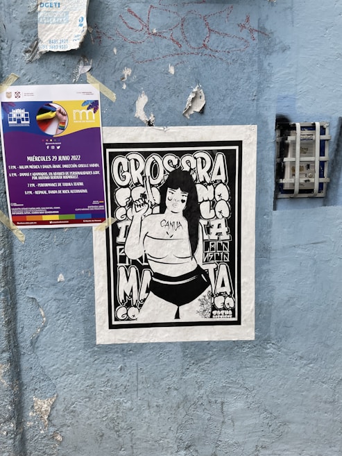 A blue wall features multiple posters. One is a colorful event poster with text about a performance scheduled for June 29, 2022. Below it, a black and white illustration of a person with long hair holding an object, surrounded by bold, graphic letters. The wall is textured and has some graffiti and peeling sections.