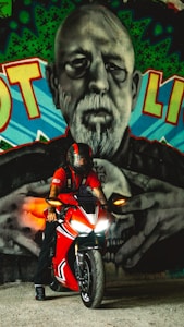 A person wearing a helmet sits on a red motorcycle, positioned in front of a large, detailed mural of a man's face. The artwork incorporates a mix of colors including green, blue, and red, with the man depicted in black and white. The motorcycle's headlights are on, casting light and creating a contrast against the vibrant graffiti.