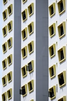 A tall building facade featuring multiple rows of uniformly square windows, each with a yellow frame. Some windows are open, creating a pattern against the white exterior walls. The architectural design highlights symmetry and contrast between the yellow frames and the white surface.