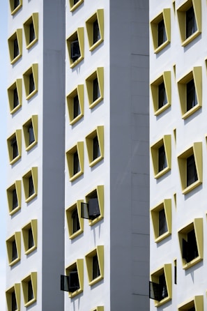 A tall building facade featuring multiple rows of uniformly square windows, each with a yellow frame. Some windows are open, creating a pattern against the white exterior walls. The architectural design highlights symmetry and contrast between the yellow frames and the white surface.