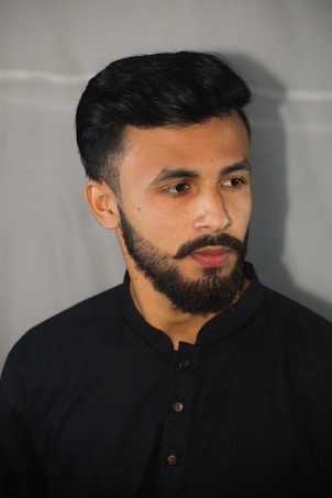 A man with a well-groomed beard and mustache, wearing a dark buttoned shirt. His hair is neatly styled, and he gazes slightly off-camera with a neutral expression.