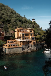 A large villa with yellow and terracotta tones sits alongside a clear, turquoise body of water. The multi-story building has various arches, balconies, and terraces, surrounded by lush green hills. People can be seen engaging in water activities, including a small boat near the villa. A smaller building with a tower is visible in the background amidst the greenery.