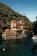 A large villa with yellow and terracotta tones sits alongside a clear, turquoise body of water. The multi-story building has various arches, balconies, and terraces, surrounded by lush green hills. People can be seen engaging in water activities, including a small boat near the villa. A smaller building with a tower is visible in the background amidst the greenery.