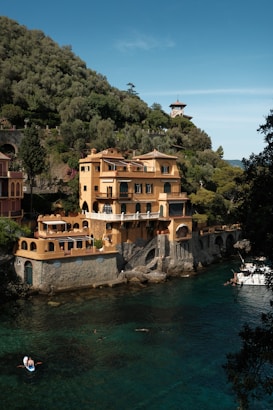 A large villa with yellow and terracotta tones sits alongside a clear, turquoise body of water. The multi-story building has various arches, balconies, and terraces, surrounded by lush green hills. People can be seen engaging in water activities, including a small boat near the villa. A smaller building with a tower is visible in the background amidst the greenery.