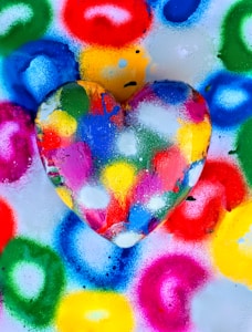 A colorful heart-shaped object is surrounded by vibrant, circular spray paint patterns on a surface. The heart is painted in various bold colors, including red, blue, green, yellow, pink, and white, creating a lively and artistic composition.
