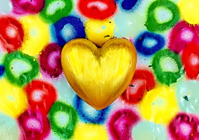 A golden heart is placed against a vibrant background of multicolored circles. The circles are spray painted in hues of red, green, blue, yellow, and purple, creating a dynamic and lively pattern.