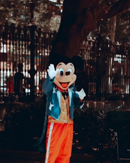 A person is dressed in a costume resembling a famous cartoon character. The costume includes a large mask with oversized ears and a cheerful expression. The outfit consists of a blue coat with star patterns, a golden vest, and bright red pants with white stripes. The background features a wrought iron fence and lush greenery, with a few silhouettes visible behind.