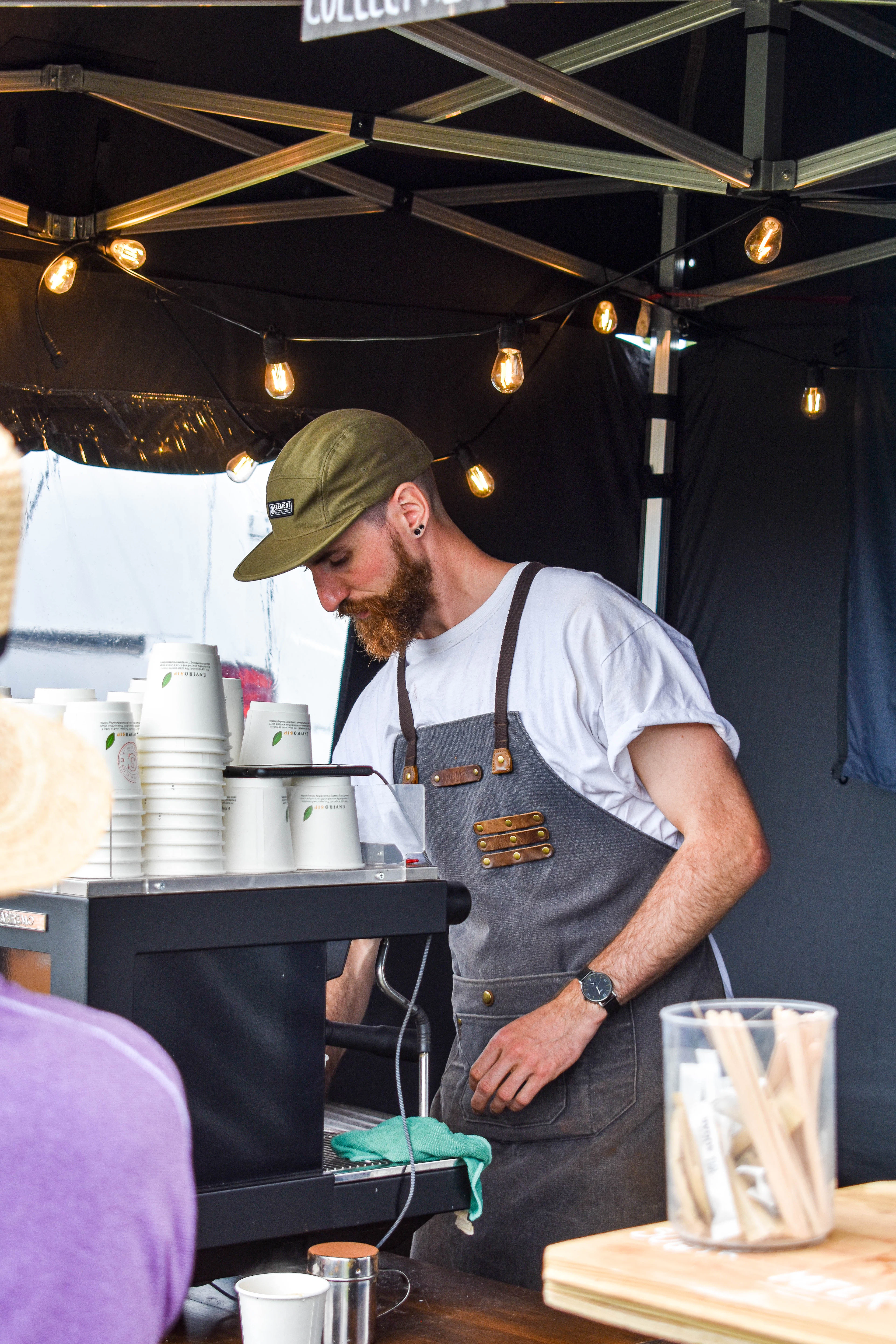 Barista in an apron working at a market coffee stall, stacks of takeaway cups beside the machine