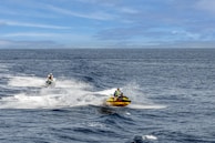 Two jet skiers navigate through the open sea, creating trails of white splashes against the deep blue water. The sky is clear with a few clouds, enhancing the expansive feel of the scene. The jet skiers wear life jackets and helmets, emphasizing safety during their adventurous ride.
