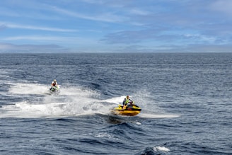 Two jet skiers navigate through the open sea, creating trails of white splashes against the deep blue water. The sky is clear with a few clouds, enhancing the expansive feel of the scene. The jet skiers wear life jackets and helmets, emphasizing safety during their adventurous ride.