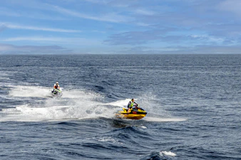 Riders laughing and speeding across the ocean on jet skis, water splashing around them.