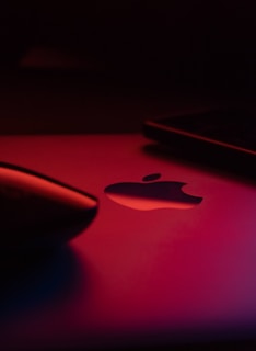 A close-up view of a metallic surface featuring a prominent cut-out logo of an apple with a smooth, reflective finish. The lighting is dim with a warm, reddish tint bathing the scene, casting soft shadows on the surface. In the foreground, the edge of a sleek, black curved object is visible, adding to the modern and sophisticated atmosphere.