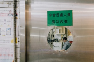 A person is seen through a circular window on a metallic door. The person appears to be seated and working in a room filled with various items like machines, calendars, and papers. There is a green sign with text above the circular window. To the left side, there are advertisements or notices posted on the wall.