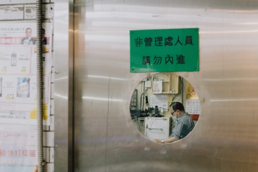 A person is seen through a circular window on a metallic door. The person appears to be seated and working in a room filled with various items like machines, calendars, and papers. There is a green sign with text above the circular window. To the left side, there are advertisements or notices posted on the wall.