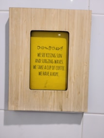 A wooden frame surrounds a yellow poster with a motivational message about hope and coffee. The text includes small icons of a cup and other food items.
