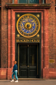 A woman walks past a large ornate door featuring a decorative clock with zodiac symbols and a sun motif. The building exterior is constructed with reddish-brown bricks, framing the entrance with intricate designs. The area above the door is inscribed with the words 'Bracken House'.