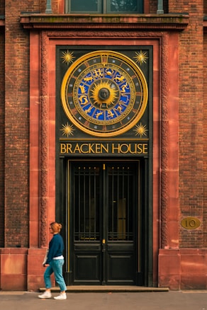 A woman walks past a large ornate door featuring a decorative clock with zodiac symbols and a sun motif. The building exterior is constructed with reddish-brown bricks, framing the entrance with intricate designs. The area above the door is inscribed with the words 'Bracken House'.