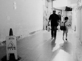 A black and white photograph capturing the rear view of a man holding a child’s hand, walking down a narrow corridor. A 'Caution Wet Floor' sign is visible on the left, with its shadow cast on the tiled floor. The walls appear worn and slightly grimy, adding an urban, gritty feel to the scene.