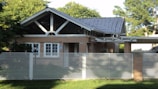 A small, single-story house with a gabled roof and large front windows is surrounded by lush greenery. The house is built with light brown bricks and features a modern design with a metal fence in the foreground. The roof appears to be made of dark gray tiles, and there is a veranda extension on one side.