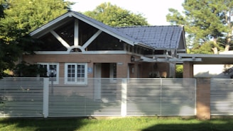 A small, single-story house with a gabled roof and large front windows is surrounded by lush greenery. The house is built with light brown bricks and features a modern design with a metal fence in the foreground. The roof appears to be made of dark gray tiles, and there is a veranda extension on one side.