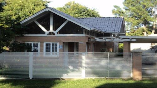 A small, single-story house with a gabled roof and large front windows is surrounded by lush greenery. The house is built with light brown bricks and features a modern design with a metal fence in the foreground. The roof appears to be made of dark gray tiles, and there is a veranda extension on one side.