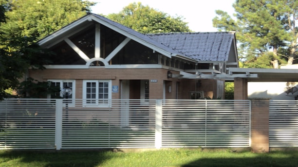 A small, single-story house with a gabled roof and large front windows is surrounded by lush greenery. The house is built with light brown bricks and features a modern design with a metal fence in the foreground. The roof appears to be made of dark gray tiles, and there is a veranda extension on one side.