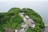 An aerial view of a lush green hill overlooking the sea. Numerous buildings with various roof types are nestled among the dense foliage, suggesting a secluded settlement or retreat. The surrounding area is heavily wooded, with vibrant greenery dominating the landscape.