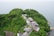 An aerial view of a lush green hill overlooking the sea. Numerous buildings with various roof types are nestled among the dense foliage, suggesting a secluded settlement or retreat. The surrounding area is heavily wooded, with vibrant greenery dominating the landscape.