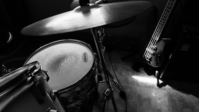A black and white image depicting a drum set and a guitar leaning against an amplifier. The focus is on a cymbal, snare drum, and the neck of a bass guitar. The setting appears to be a music studio or practice room with tiled flooring and soft lighting creating a dramatic effect.