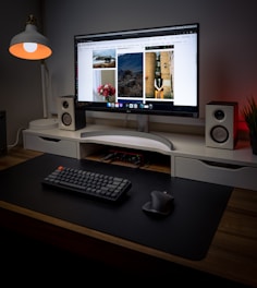 A modern, well-organized desktop setup featuring a large monitor displaying a gallery of images. The desk includes a minimalistic keyboard and ergonomic mouse on a black mat. On either side of the monitor are two white speakers. A stylish lamp with a warm bulb is on one side, providing ambient lighting. The setting is accentuated with a clean and minimal design aesthetic, with an additional plant adding a touch of nature.