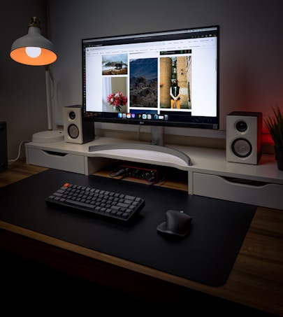 A modern, well-organized desktop setup featuring a large monitor displaying a gallery of images. The desk includes a minimalistic keyboard and ergonomic mouse on a black mat. On either side of the monitor are two white speakers. A stylish lamp with a warm bulb is on one side, providing ambient lighting. The setting is accentuated with a clean and minimal design aesthetic, with an additional plant adding a touch of nature.