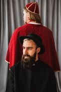 A man with a beard is wearing a black hat and clerical attire, gazing off to the side. Behind him, another person with long hair is dressed in a red robe and hat, facing away. A gray curtain serves as the backdrop.