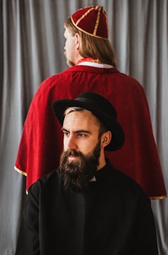 A man with a beard is wearing a black hat and clerical attire, gazing off to the side. Behind him, another person with long hair is dressed in a red robe and hat, facing away. A gray curtain serves as the backdrop.