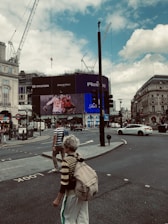 An urban street scene with a large electronic billboard displaying various advertisements, including a Coca-Cola ad. Construction cranes are visible against a partly cloudy sky. People cross the street, and vehicles are present, including a white car. The setting appears to be a busy city square with buildings and traffic signs.