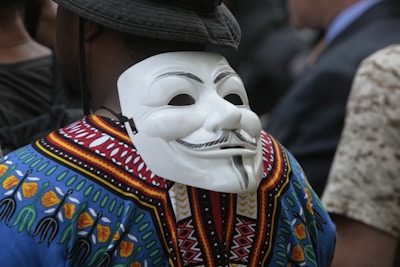A person wearing a colorful, patterned shirt and a black hat has a white mask with a stylized mustached face attached to the back of their head. The shirt features vibrant designs with blue, yellow, red, and green colors, resembling traditional patterns.