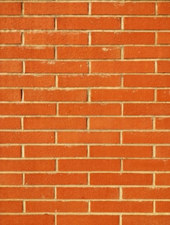 A wall constructed from red bricks, each uniformly sized and horizontally arranged in staggered rows with beige mortar filling the gaps between the bricks. The surface appears textured and the color is a vivid, warm red.