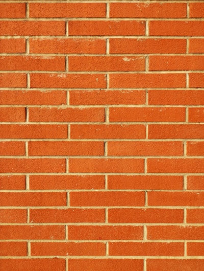 A wall constructed from red bricks, each uniformly sized and horizontally arranged in staggered rows with beige mortar filling the gaps between the bricks. The surface appears textured and the color is a vivid, warm red.