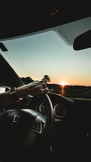 A driver's hand is on the steering wheel of a Honda car, with the dashboard and speedometer visible. The sun is setting on the horizon, casting a warm glow over the scenery and creating a silhouette of trees and distant hills.