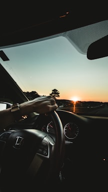A driver's hand is on the steering wheel of a Honda car, with the dashboard and speedometer visible. The sun is setting on the horizon, casting a warm glow over the scenery and creating a silhouette of trees and distant hills.