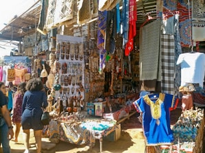 A bustling market stall filled with a variety of colorful textiles, traditional masks, and handcrafted items. Vibrant tapestries and garments hang alongside shelves and tables displaying an array of intricate wooden carvings and souvenirs. Several people are browsing the items, contributing to the lively atmosphere.
