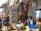 A bustling market stall filled with a variety of colorful textiles, traditional masks, and handcrafted items. Vibrant tapestries and garments hang alongside shelves and tables displaying an array of intricate wooden carvings and souvenirs. Several people are browsing the items, contributing to the lively atmosphere.