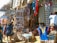 A bustling market stall filled with a variety of colorful textiles, traditional masks, and handcrafted items. Vibrant tapestries and garments hang alongside shelves and tables displaying an array of intricate wooden carvings and souvenirs. Several people are browsing the items, contributing to the lively atmosphere.
