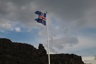 A flagpole with the Icelandic flag flutters against a backdrop of a cloudy sky. Below the flag, there is a rugged, dark rocky landscape.