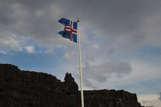 A flagpole with the Icelandic flag flutters against a backdrop of a cloudy sky. Below the flag, there is a rugged, dark rocky landscape.