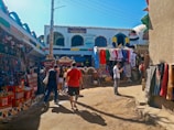 A bustling outdoor market scene with colorful textiles draped on the walls and hanging from stalls. Several people are walking and browsing through the vibrant market area filled with garments and traditional crafts. The buildings are adorned with bright and intricate patterns, adding to the lively and festive atmosphere.