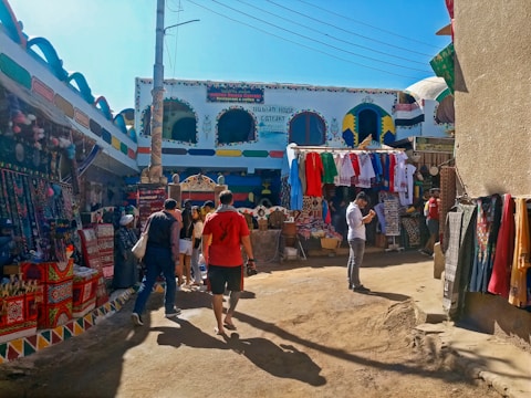 A bustling outdoor market scene with colorful textiles draped on the walls and hanging from stalls. Several people are walking and browsing through the vibrant market area filled with garments and traditional crafts. The buildings are adorned with bright and intricate patterns, adding to the lively and festive atmosphere.