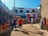 A bustling outdoor market scene with colorful textiles draped on the walls and hanging from stalls. Several people are walking and browsing through the vibrant market area filled with garments and traditional crafts. The buildings are adorned with bright and intricate patterns, adding to the lively and festive atmosphere.