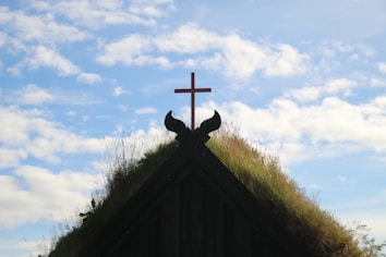 A grass-covered roof of a building is topped with a wooden cross, nestled between decorative wooden elements that resemble traditional Scandinavian architecture under a blue sky with scattered clouds.