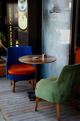 A cozy corner with a small round wooden table flanked by two plush chairs, one in green and the other in blue with an orange seat. The setup is on a wooden floor, next to a window with a frosted design, creating a relaxed atmosphere. Nearby, there is a decorative plant and a wall with food images and text in yellow and white.