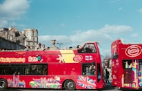 A brightly colored red double-decker bus with 'City Sightseeing Bath' written on the side. The bus features various vibrant designs, including the Union Jack and iconic city landmarks. People are seated on the upper open-top deck, enjoying the view of historical buildings and a clear sky.